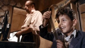Chłopiec z uśmiechem ogląda taśmę filmową w kabinie projekcyjnej. W tle stoi starszy mężczyzna przy projektorze.