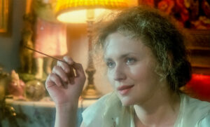 kadr z filmu. dojrzała blondynka z kręconymi włosami i papierosem w ręce uśmiecha się zawadiacko. W tle lampa z abażurem i wnętrze pokoju w stylu retro.