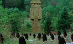 na tle drzew na polanie figura - ogromna postać w kształcie człowieka. poniżej zgromadzone stwory z bardzo długimi ciemnymi włosami zakrywającymi ciało