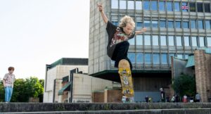 na tle biurowca, osoba z półdługimi blond włosami robi wyskok na deskorolce