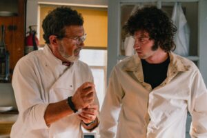 Kadr z filmu „Tony”: Dwóch kucharzy rozmawia w kuchni. Starszy mężczyzna w białym stroju kucharskim zwraca się do młodszego, kręconowłosego mężczyzny.