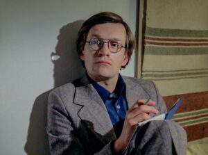 mężczyzna w okularach, szarym garniturze, party o ścianę, na której wisi koc, trzyma notatnik i długopis